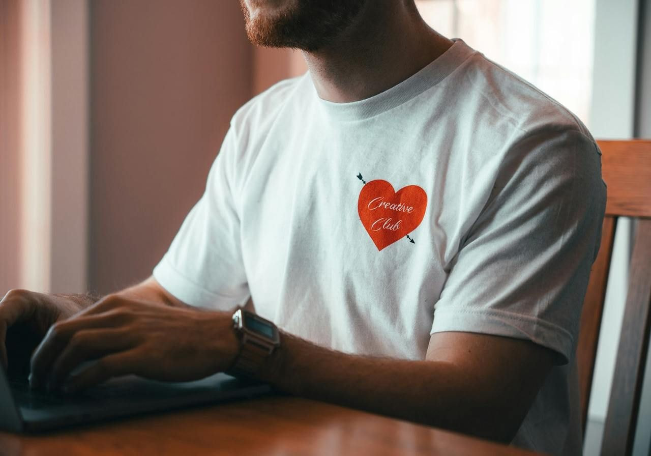 A man is sitting by a laptop, wearing a white t-shirt with a heart print.