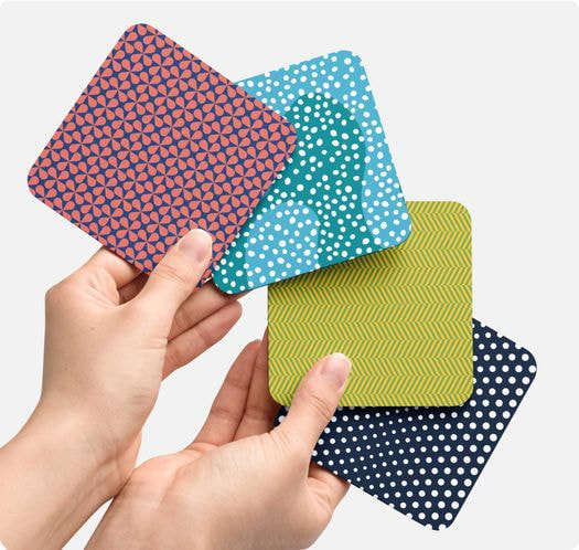 Two hands are holding customized coasters with abstract designs of pink, blue, green, and dark blue color